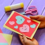 Child's hands making a Valentine's Day card with cut-out paper hearts in pink and red, surrounded by scissors, glue, and colored paper on a purple surface.