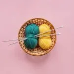 Top view of a woven basket containing balls of teal and yellow yarn with knitting needles on a pink background.