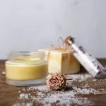 Homemade body care products including jars of yellow body butter and a glass tube of bath salts, with a dried rose on a wooden surface.