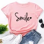 Flatlay of a pink t-shirt with 'Smile' text, paired with jeans, white sneakers, sunglasses, and a plant