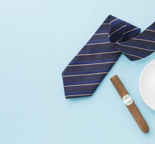 Navy striped tie, white coffee cup, and cigar arranged on light blue background, suggesting a Father's Day gift