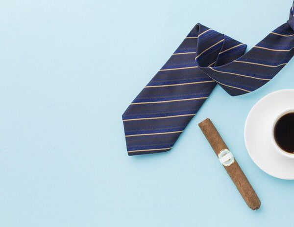 Navy striped tie, white coffee cup, and cigar arranged on light blue background, suggesting a Father's Day gift