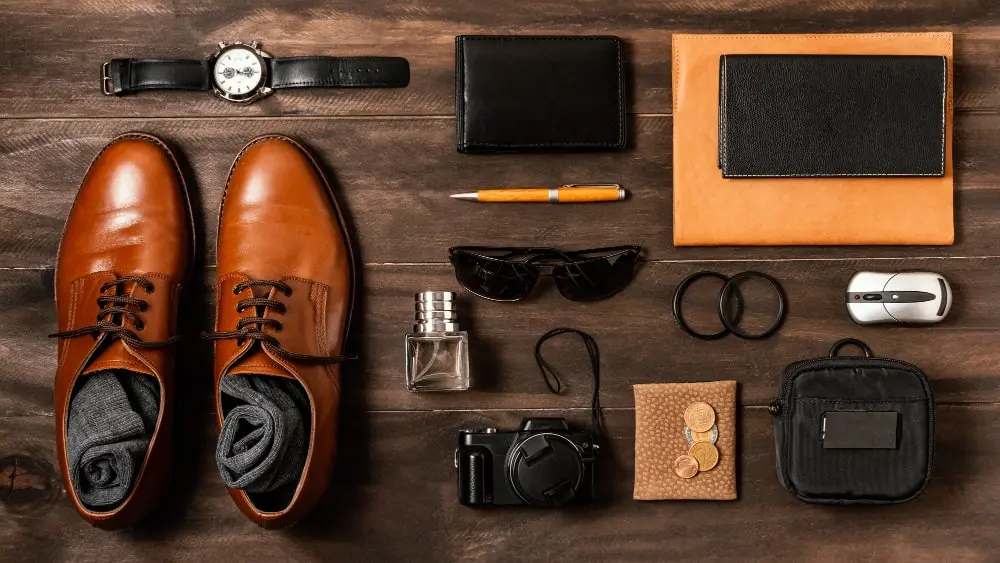 Flatlay of men's accessories on wooden background including brown leather shoes, watch, wallet, sunglasses, camera, perfume bottle, notebook, pen, computer mouse, and various small items for business and travel.