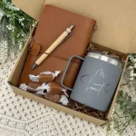 Sustainable gift box containing a brown leather journal, wooden pen, gray personalized tumbler with 'M Smith' engraved, wrapped caramels, and decorative greenery on a textured white background.